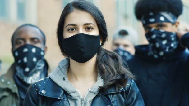 Close up of young Caucasian beautiful woman in mask looking straight at camera outdoor at street riot. Portrait of girl with male Afrcan Americans on background. Female protester at manifestation. - Footage, Video