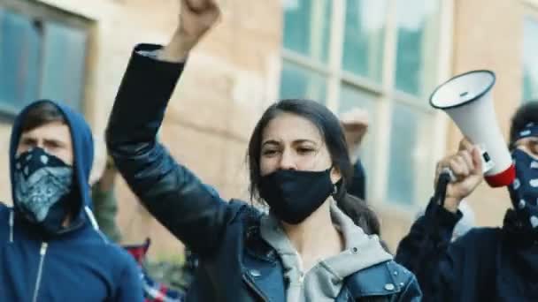 Красивая белая молодая женщина в маске кричит лозунги в знак протеста в многонациональной мужской толпе. Женщина довольно протестует против проявления прав человека и против полицейского насилия, выкрикивая лозунги
. - Кадры, видео