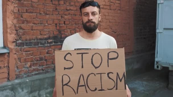 Brodaty biały mężczyzna w masce stoi pod czerwoną ścianą z kartonowym plakatem w ręku z napisem - STOP RACISM. Protest pojedynczy. - Materiał filmowy, wideo