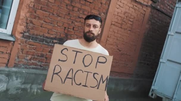 L'uomo caucasico barbuto in maschera sta contro parete rossa con manifesto di cartone in mano con iscrizione - STOP RACISM. Protesta unica. - Filmati, video