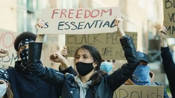 Mooie blanke jonge vrouw in masker hoding poster Vrijheid essentieel en schreeuwen motto 's bij protest in multi-etnische menigte. Vrouwelijke demonstrant bij manifestatie voor mensenrechten en tegen geweld. - Video