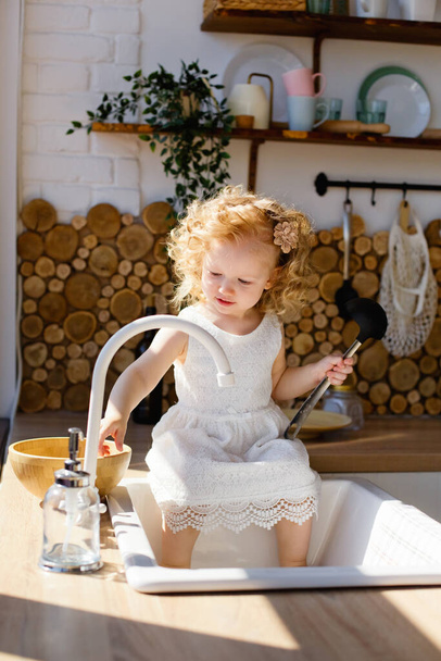 A little girl is sitting on a table in a bright Scandinavian-style kitchen. Playing with dishes, cereals, beans. The concept of children's development Montessori. - Foto, imagen