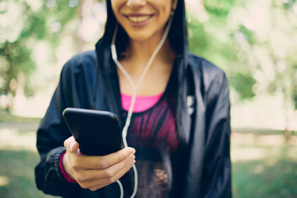 Closeup of smiling caucasian female runner standing in nature and using smart phone. - 写真・画像