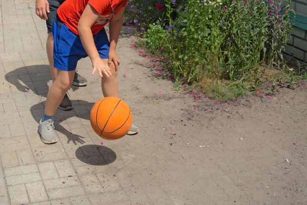 Adolescente juega baloncesto con su padre. Vista de cerca sin rostros. Chico y hombre rebotan la pelota en un patio trasero. Los aficionados al deporte en el soleado día de verano
 - Foto, Imagen