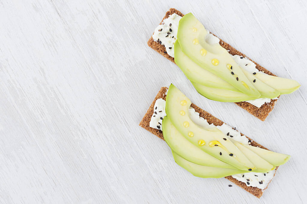 Fresh summer healthy appetizer of whole grain rye crisps breads with avocado slices, olive oil, black sesame seeds, cream cheese on white wood board, top view, copy space, closeup. - Zdjęcie, obraz