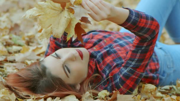 Portret pięknej kobiety z czerwonymi włosami leżącej w żółtych jesiennych liściach. Kobieta model korzystających z życia na zewnątrz natura tło 4k zwolnione tempo - Materiał filmowy, wideo