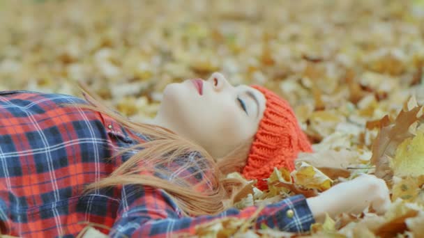 Sarı sonbahar yapraklarında yatan kırmızı şapkalı güzel bir kadının portresi. Kadın model açık havada hayatın tadını çıkarıyor. Arka plan 4k ağır çekim. - Video, Çekim