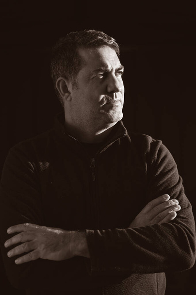 Уверенный взрослый мужчина, стоящий в темноте и смотрящий в сторону от камеры на черном фоне
. - Фото, изображение