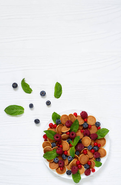 Gezond zomerontbijt met kleine pannenkoeken, verse bessen en muntblaadjes bovenaanzicht op witte houten tafel - Foto, afbeelding