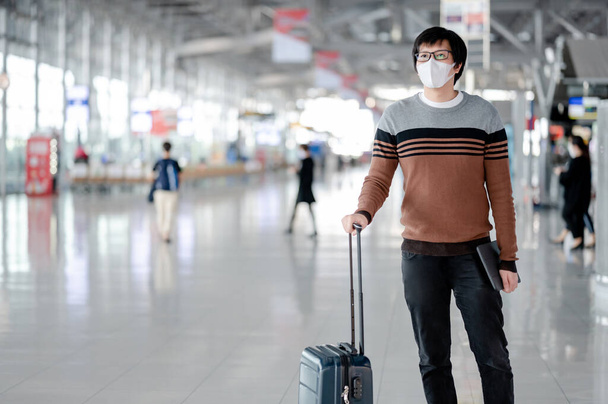 Asiático turista vestindo máscara facial transportando mala de bagagem à espera de check-in no terminal do aeroporto. Prevenção da pandemia de Coronavirus (COVID-19) quando viajar. Consciência da saúde e distanciamento social
 - Foto, Imagem
