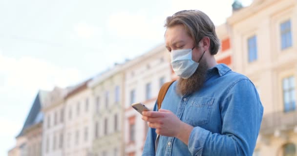 Hombre caucásico con barba y máscara médica de pie en la calle y mensaje de texto en el teléfono móvil. Hombre en la protección respiratoria es al aire libre y golpeando o desplazándose en el teléfono inteligente. Navegar en línea
. - Metraje, vídeo