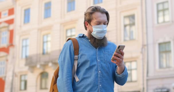 Fehér férfi szakállal és orvosi maszkkal az utcán áll és üzenetet ír a mobilján. Férfi a légúti védelem a szabadban, és koppintás vagy görgetés okostelefonon. Böngészés online. - Felvétel, videó