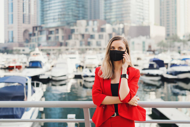 Kırmızı takım elbiseli, yeniden kullanılabilir siyah maske takan genç bir kadın şehir merkezinde duruyor ve akıllı telefondan arama yapıyor. Covid 19 ya da koronavirüs salgını sırasında kadın girişimci - Fotoğraf, Görsel