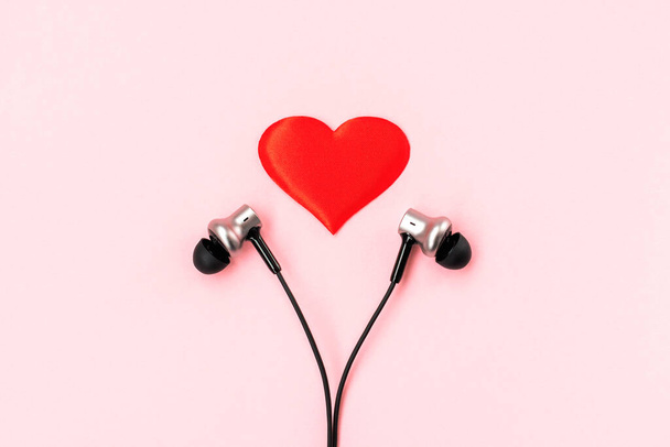Corazón rojo con un par de auriculares de música negra para un teléfono inteligente sobre un fondo rosa pastel. Concepto romántico y amoroso. Bonita postal de regalo. Redacción plana minimalista. Vista superior. Primer plano.
. - Foto, Imagen
