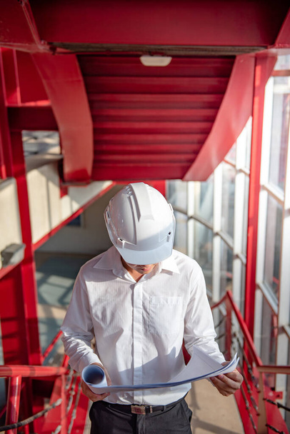 Azjatycki inżynier budowlany lub pracownik budowlany noszący ochronny hełm, trzymający plany. Mężczyzna architekt pracujący na czerwonych schodach na placu budowy. Koncepcje projektowania budynków i architektury - Zdjęcie, obraz