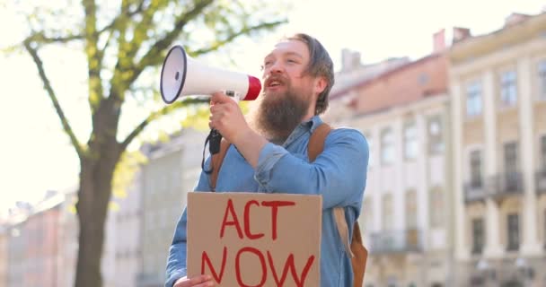 Белый мужчина-активист с бородой, говорящий и кричащий в громкоговорителе. Мужчина держит плакат "Закон сейчас" на политической или экологической одинокой демонстрации. Единый протест на городской площади
. - Кадры, видео