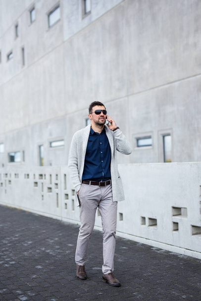 портрет красивого бизнесмена, ходящего и разговаривающего по смартфону на фоне современного здания
 - Фото, изображение