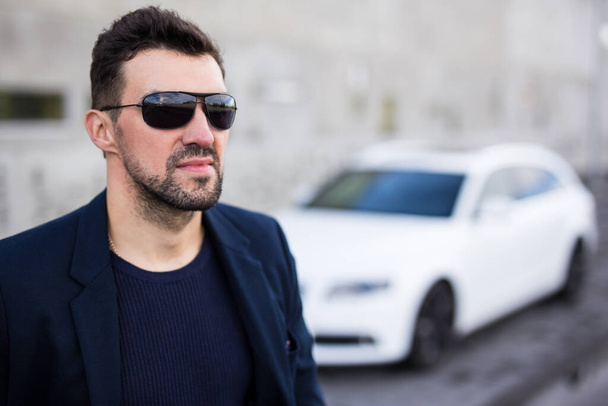 close-up portret van knappe zakenman in zonnebril poseren met zijn witte auto - Foto, afbeelding