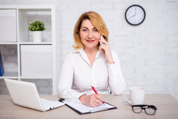 cheerful mature business woman sitting in modern office, talking by phone and writing something on clipboard - Foto, Imagen