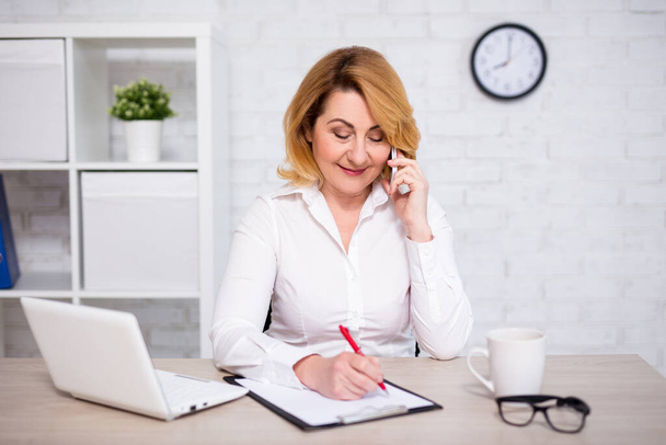 portret van volwassen zakenvrouw zitten in modern kantoor, praten via de telefoon en het schrijven van iets op klembord - Foto, afbeelding