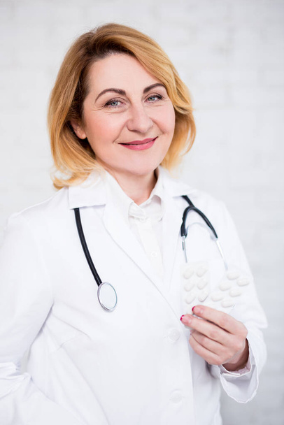 health care and pharmacy concept - portrait of mature female doctor holding pills over white wall - Photo, Image