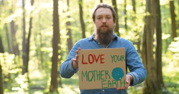 Portrait d'un jeune éco-aciviste caucasien debout dans un parc vert ensoleillé et tenant la table avec des mots Love our Mother Earth. Bel homme avec une déclaration pour une nature propre et sûre. - Séquence, vidéo