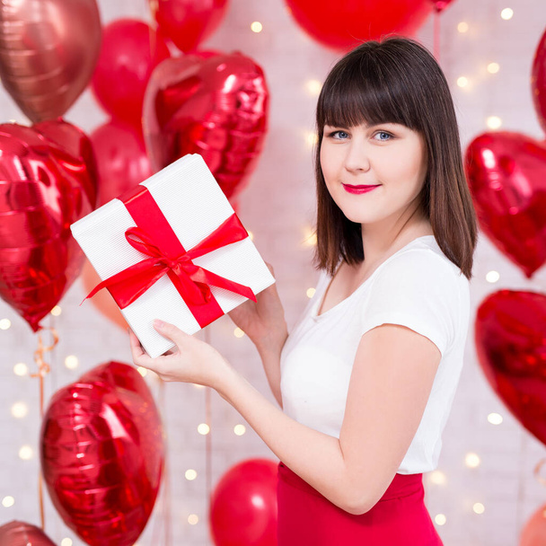 valentine's day concept - beautiful woman holding gift box over red heart-shaped balloons background - Fotoğraf, Görsel