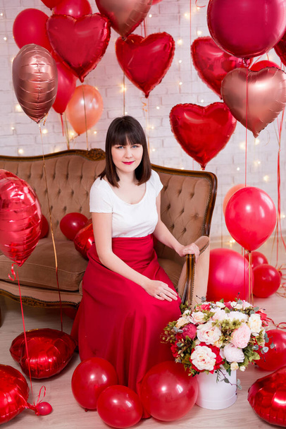 Sevgililer Günü konsepti. Kırmızı kalp şeklinde helyum balonları olan eski bir koltukta oturan genç bir kadının tam boy portresi. - Fotoğraf, Görsel