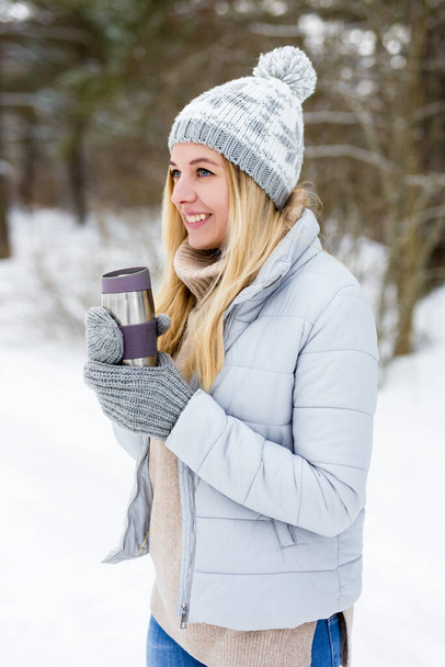 portrait of young beautiful blond woman with thermo cup of coffee in winter park or forest - Photo, Image