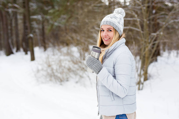 młoda piękna blondynka spacerująca w zimowym parku lub lesie z termo filiżanką kawy - Zdjęcie, obraz