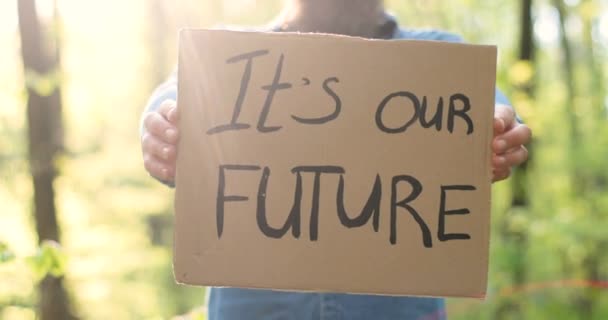Close-up van karton poster met woorden Het is onze toekomst in handen van de Kaukasische man. Mannelijke eco-activist staat in bos of park op zonnige dag met eenzaam protest. - Video