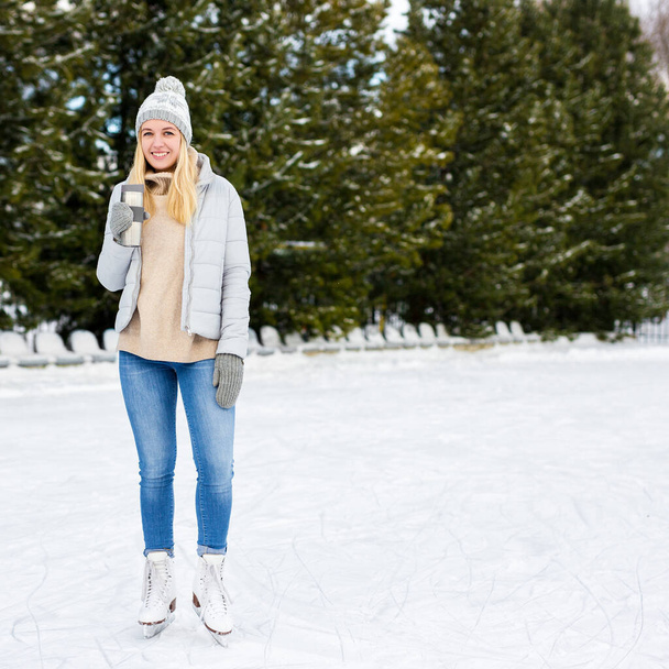 full length portrait of happy young woman ice skating and drinking coffee at rink in winter park - 写真・画像