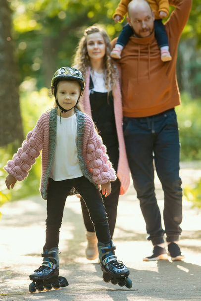 Young happy family of parents and two little kids walking in the forest - a girl skating on the rollers - Foto, Imagen