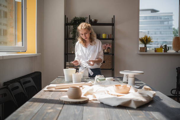 Młoda Europejka piękna garncarka w kreatywnym procesie produkcji ceramiki przy stole w warsztacie. Włosy trzepoczą na wietrze w słoneczny dzień w pracowni garncarskiej. - Zdjęcie, obraz