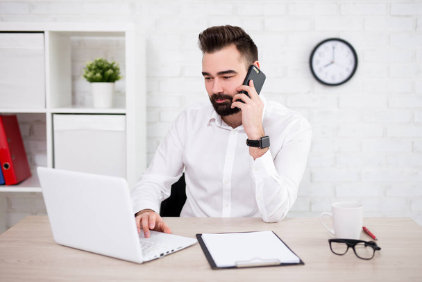 портрет красивого бородатого бизнесмена, использующего компьютер и разговаривающего по телефону в современном офисе
 - Фото, изображение