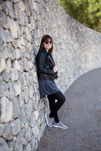 full length portrait of young woman in leather jacket and sunglasses posing over stone wall - Photo, Image
