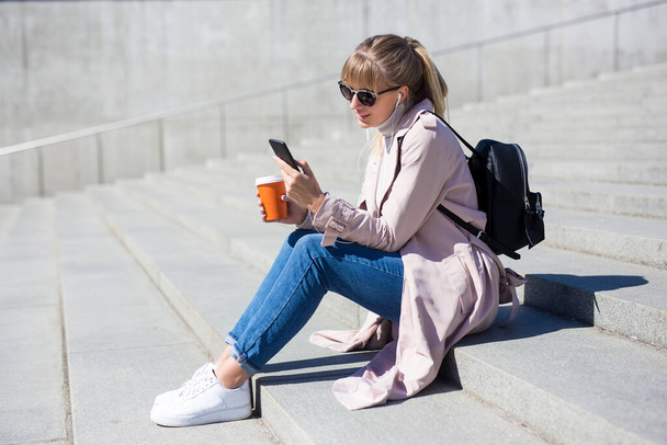 életmód és utazási koncepció - kültéri portré fiatal nő ül a lépcsőn okostelefonnal és papír csésze kávé - Fotó, kép