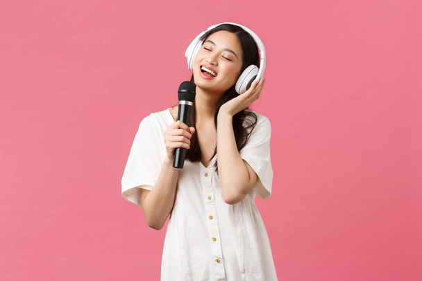 Schönheit, Menschen Emotionen und Technologie-Konzept. Sorglos glücklich asiatische Mädchen mit Handy-Karaoke-Anwendung, singen in Mikrofon, Musik hören in Kopfhörer, rosa Hintergrund - Foto, Bild