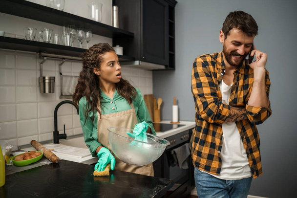 Young multiethnic couple is cleaning their kitchen. Man is lazy and woman is angry. - Photo, Image