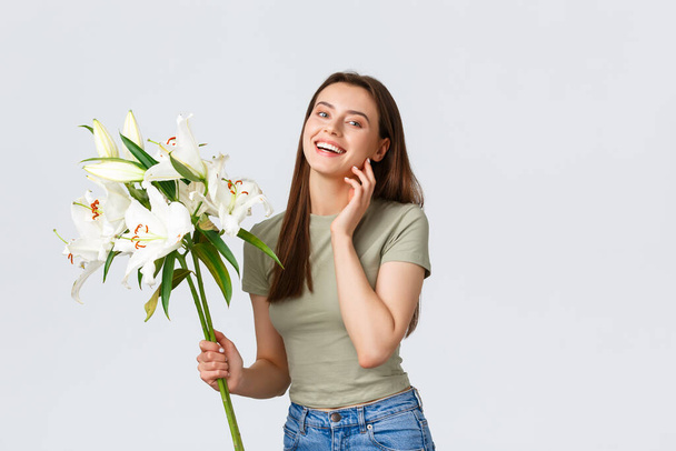 Smiling good-looking caucasian woman receive flowers delivery from florist shop, looking happy at bouquet white lilies. Girl dreamy touching leaf and thinking of boyfriend who made romantic gesture - Φωτογραφία, εικόνα