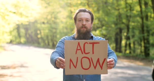 Portré kaukázusi fiatal férfi öko-acivista áll zöld napos erdőben az út mellett, és kezében poszter szavakkal Act Now. Jóképű férfi a tiszta és biztonságos természetért. Környezetvédelmi aktivitási koncepció. - Felvétel, videó