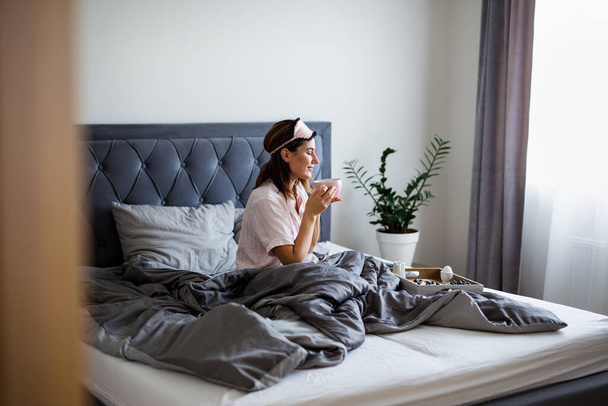 good morning concept - portret van een mooie vrouw in pyjama zittend op bed en koffie drinkend in de slaapkamer - Foto, afbeelding