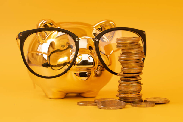 Golden piggy bank wearing glasses with money towers. Stack of euro coins near golden money box. Money pig, money saving, moneybox, pile of coins. Finance and investments. Free space for text. - Photo, Image
