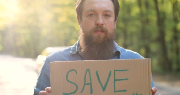 Close-up van een blanke man met een kartonnen poster met woorden Red de aarde in handen van een blanke man. Mannelijke eco-activist staat in bos of park aan de kant van de weg op zonnige dag met enkele protest. - Video