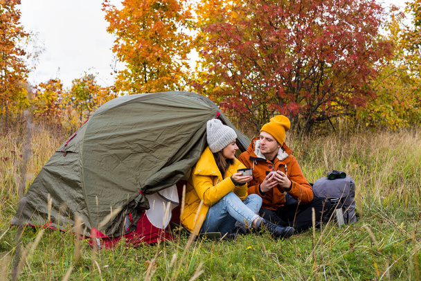 旅行、トレッキング、ハイキングのコンセプト-幸せな若いカップルは秋の森の緑のテントの近くでお茶を飲む - 写真・画像
