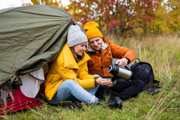 トレッキングとハイキングのコンセプト-秋の森の中で緑のテントとお茶を飲むかわいいカップルの肖像画 - 写真・画像
