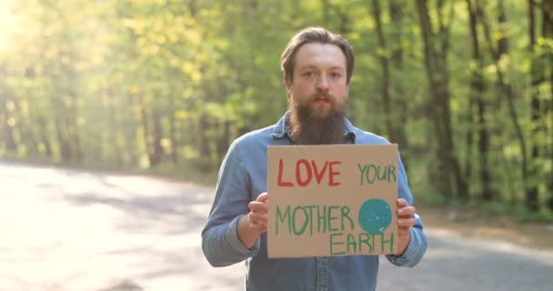 Portrait of Caucasian young male eco acivist standing in green sunny park on road side and holding table with words Love our Mother Earth. Handsome man with board statement for clean and safe nature. - Footage, Video