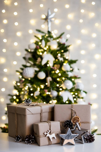 クリスマスと新年のコンセプト-ガーランドライトと白い壁の上に装飾されたクリスマスツリーの近くのベージュのギフトボックス - 写真・画像