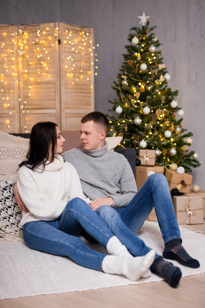 christmas, new year and love concept - young couple in love sitting in decorated living room with Christmas tree and gifts - Foto, Imagen