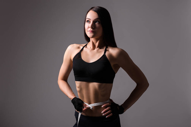 portrait of young beautiful sporty woman with muscular body measuring waistline over gray background - Photo, Image